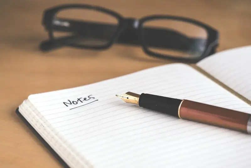 An open notebook with the word Notes written on it, a fountain pen and reading glasses